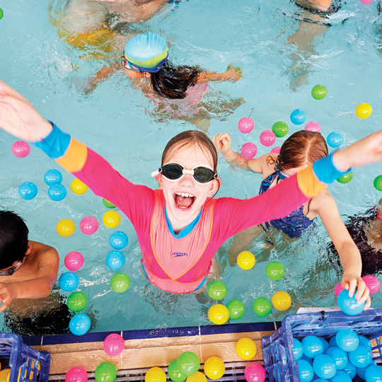 Young girl in pink rash fest and googles in a pool smiling with arms outstretched surrounded by coloured balls